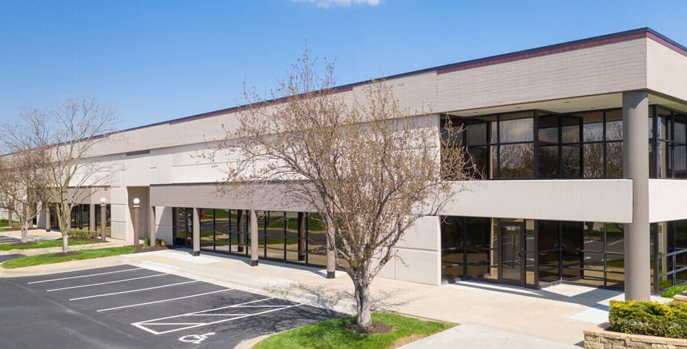 A modern, single-story office building with large glass windows, surrounded by bare trees and an empty parking lot under a clear blue sky.