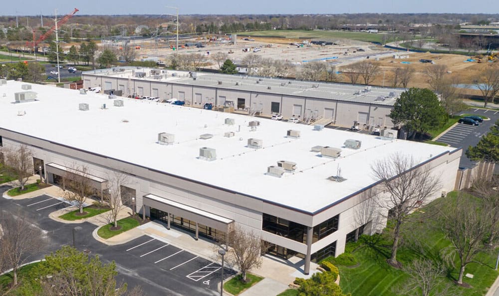 Aerial view of a large white-roofed industrial building with parking spaces. Surroundings include trees, a paved road, and a construction site in the background.