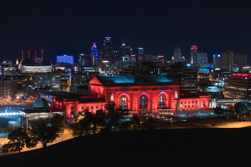 Kansas City skyline at night with an illuminated Union Station building in the foreground, glowing red, surrounded by other lit-up structures.