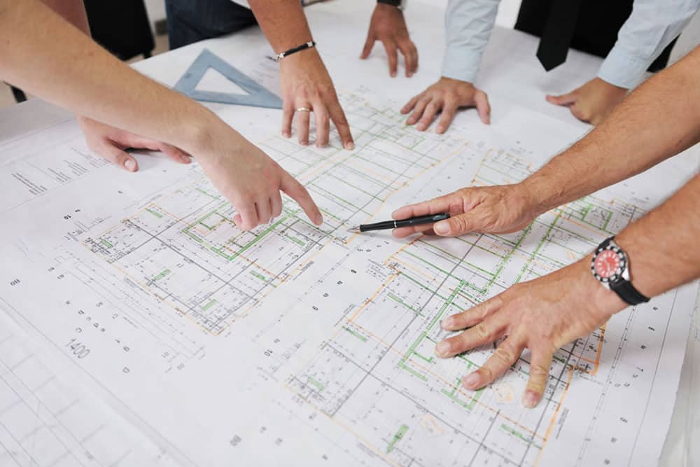 Several hands pointing at and gesturing over architectural blueprints spread out on a table.